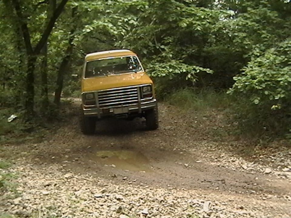 Yellow van with chrome grille driving on a dirt road through trees