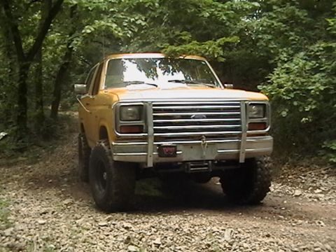 1986 Ford Bronco - This 1986 Ford Bronco is captured on a dirt path surrounded by trees, a prominent chrome grille guard.