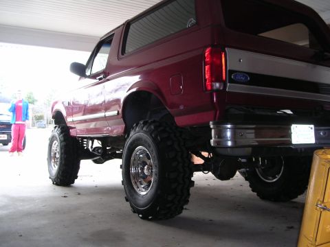 1995 Ford Bronco - A lifted red Ford Bronco with oversized tires is parked under a cover.