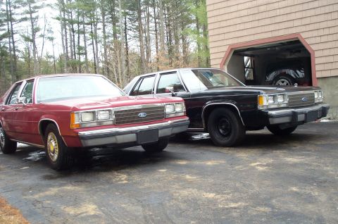 1991 Ford LTD Crown Victoria - Two Ford Crown Victoria LTD sedans, one red and one black, are parked in front of a garage. The red car is a 1991 model.