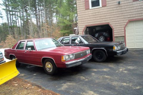 1991 Ford LTD Crown Victoria - Two sedans, a red 1991 Ford Crown Victoria LTD and a black one, are parked outside a garage.