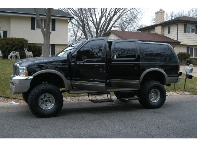 Side view of a lifted black Ford Excursion SUV with tan lower trim and large off-road tires on a suburban street