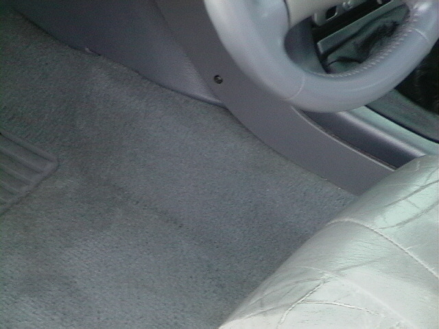 Car interior showing gray carpet, part of a steering wheel, and a quilted seat. Supermotors.net watermark visible