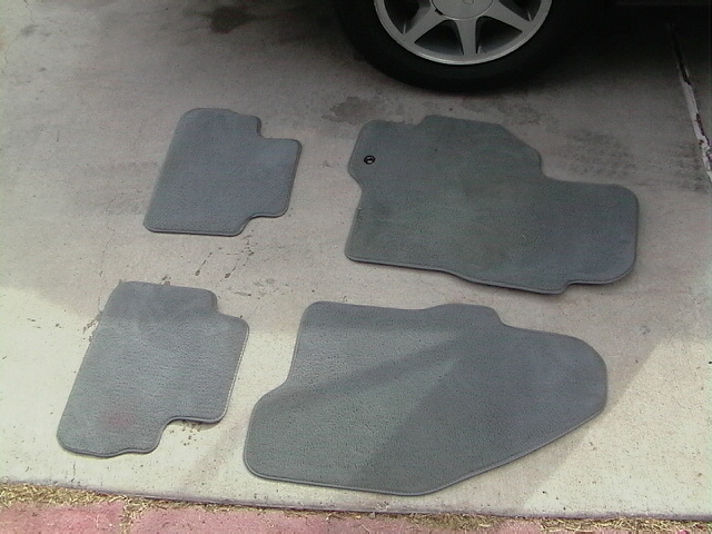 Four gray car floor mats laid out on concrete, with a car wheel visible in the upper right corner