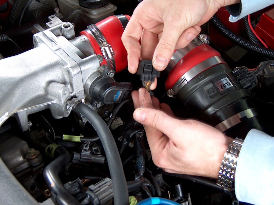 Hands back-probing an engine bay connector with a needle beside a red silicone intake hose