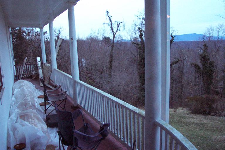 View from a covered porch with chairs and covered items, looking out at a bare-branched forest and distant hills