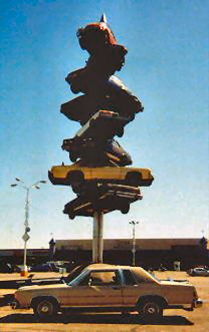 1985 Ford LTD Crown Victoria - A sculpture of stacked cars stands tall against a clear blue sky, with a tan sedan parked below.