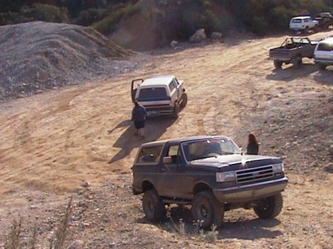 1991 Ford Bronco - Elevated view of a lifted dark Ford Bronco on mud-terrain tires parked on a dusty off-road park trail, seen from the front three-quarter wi…