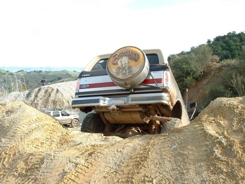 1991 Ford Bronco - Rear view of a lifted Ford Bronco climbing over a dirt mound on an off-road trail, flexed enough to expose the fuel tank, driveshaft and re…
