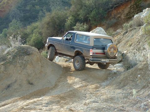1991 Ford Bronco - Blue and tan two-tone Ford Bronco flexing its lifted suspension as it crawls over a rocky dirt berm on a hillside trail, shot from the rear…