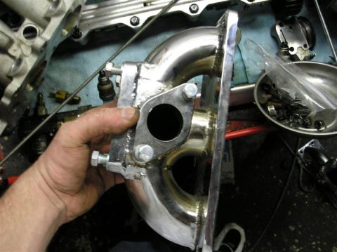 1994 Ford Probe - A custom exhaust manifold is held up, showing its polished metal and bolted flange. Other engine parts are visible in the background.
