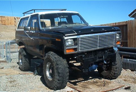 1986 Ford Bronco - This lifted 1986 Ford Bronco features a black body with a white roof and a prominent roof rack, sitting on large off-road tires.