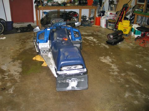 1990 Ford Mustang - A blue snowmobile sits on a wet garage floor, with various items stored on shelves behind it.