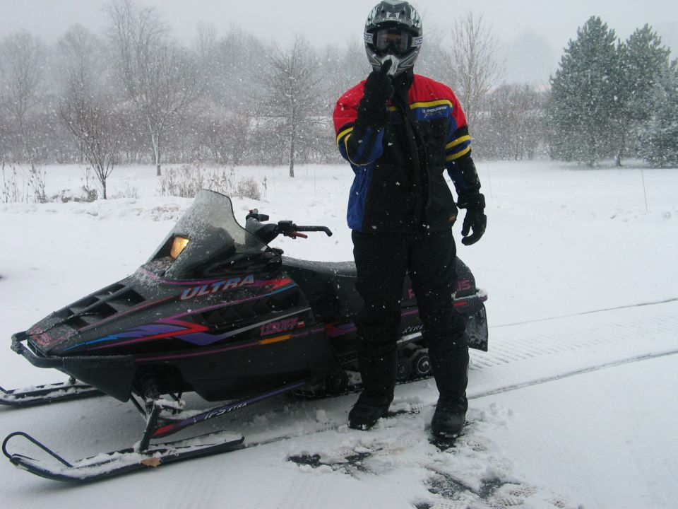 Person in snow gear stands next to a snowmobile in a snowy landscape