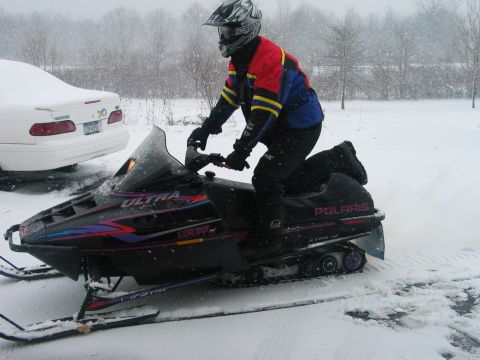 1990 Ford Mustang - A person rides a Polaris snowmobile in a snowy environment next to a white car.