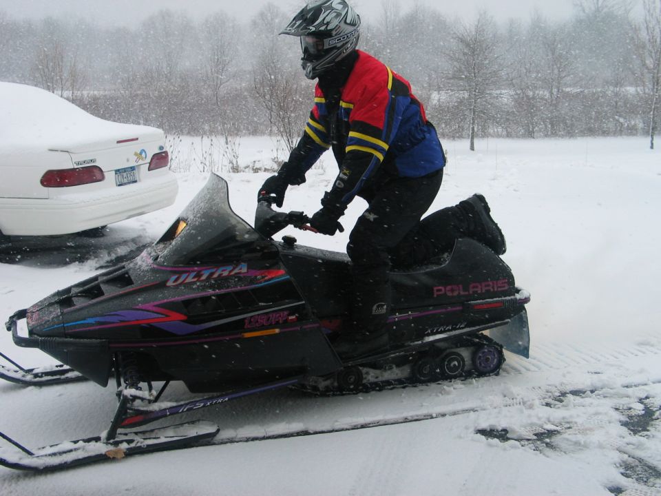Person on black Polaris snowmobile in snow, white car parked nearby