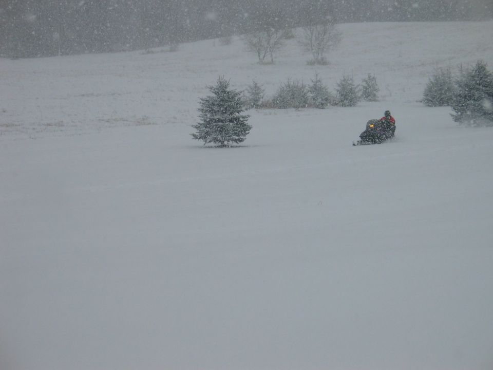 Snowmobile with rider in orange jacket moving across a snowy field during snowfall