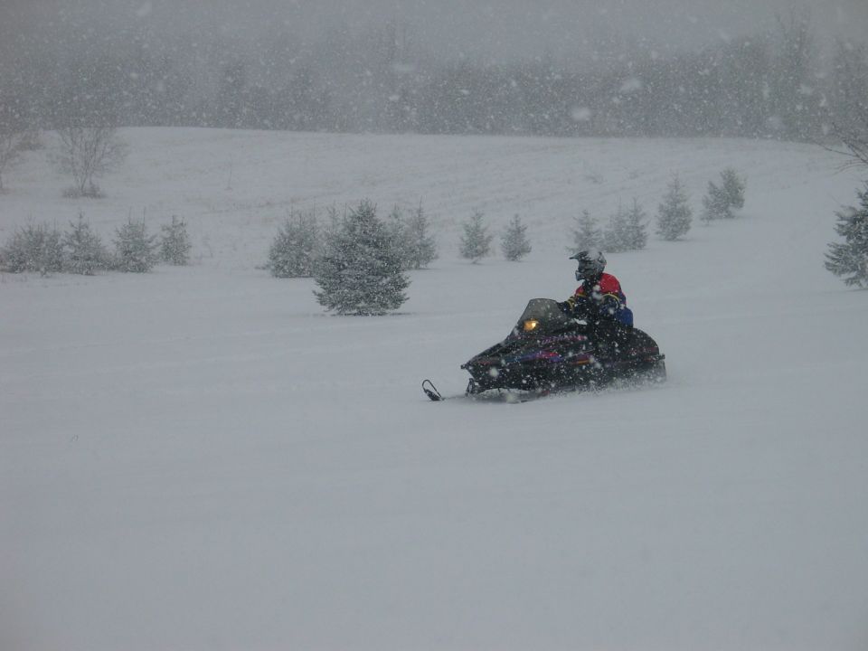 Person riding snowmobile through snowy field with trees in background