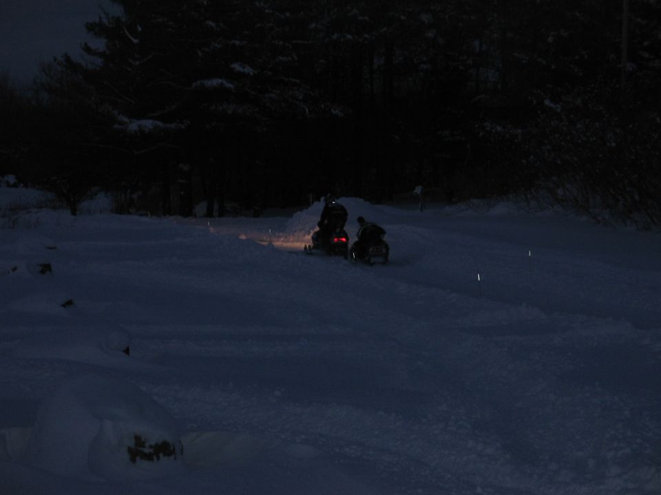 Two snowmobiles ride through a snowy landscape at night with trees in the background