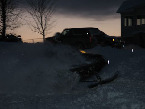 1990 Ford Mustang - A snowmobile is partially submerged in snow, with two SUVs visible in the background on a winter evening.