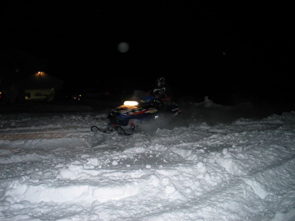 Snowmobile kicking up snow at night, with a building in the background