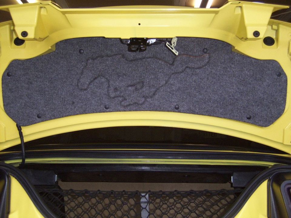 Yellow trunk interior with gray carpeted panel showing embossed running horse logo. Visible latch and wiring