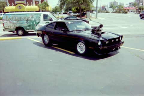 1978 Ford Mustang II - A black modified coupe with a prominent supercharger is parked in a lot next to a van.