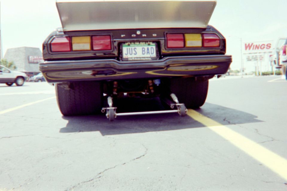 Rear view of a drag car with a large spoiler and wide tires. License plate reads 'JUS BAD'