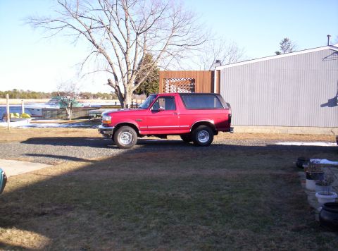 1995 Ford Bronco - A red 1995 Ford Bronco is parked in a yard with a body of water and trees in the background.
