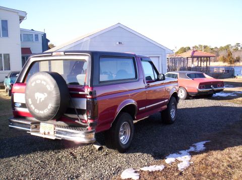 1995 Ford Bronco - A red Ford Bronco is parked in a gravel lot next to a vintage muscle car, with a house and garage in the background.