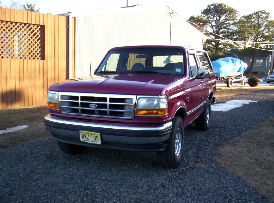 Maroon Ford Bronco parked on gravel lot, wooden fence behind it. Boat on trailer in background