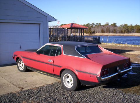 1971 Ford Mustang - A red 1973 Ford Mustang coupe with a black vinyl roof is parked outside a garage next to a body of water.