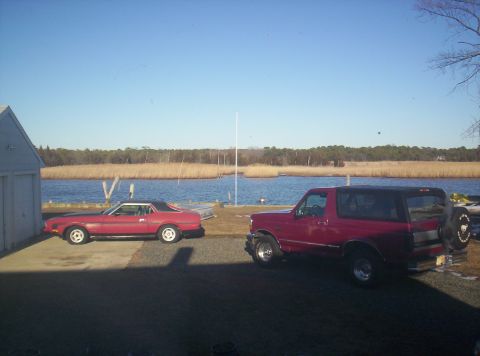 1971 Ford Mustang - Two vehicles, a red coupe and a red SUV, are parked near a body of water on a sunny day.