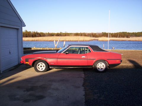 1971 Ford Mustang - Side view of a red 1971 Ford Mustang Grande coupe, a black vinyl roof and parked next to a garage with a body of water in the background.