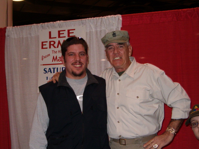 Man in vest and man in cap pose for photo in front of red curtain with sign that says 'LEE ERN... from Ma... SATUR... 1:'