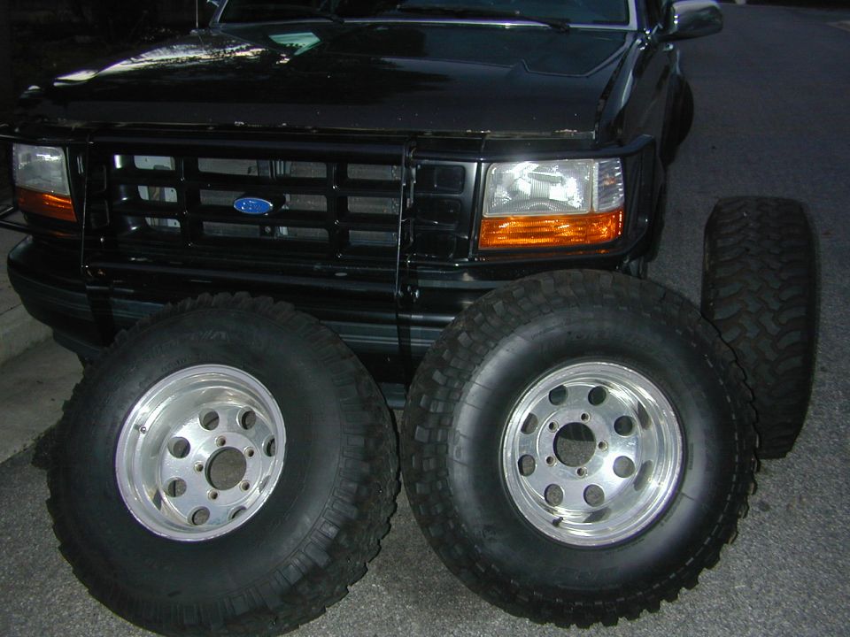 Black Ford Bronco front end with three off-road tires on polished aluminum wheels leaning against it