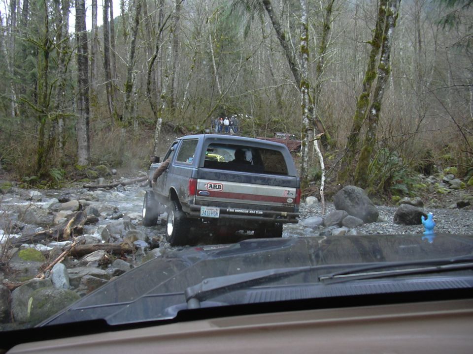 Gray and red Ford Bronco crossing a rocky creek, seen through a windshield with a Cookie Monster toy