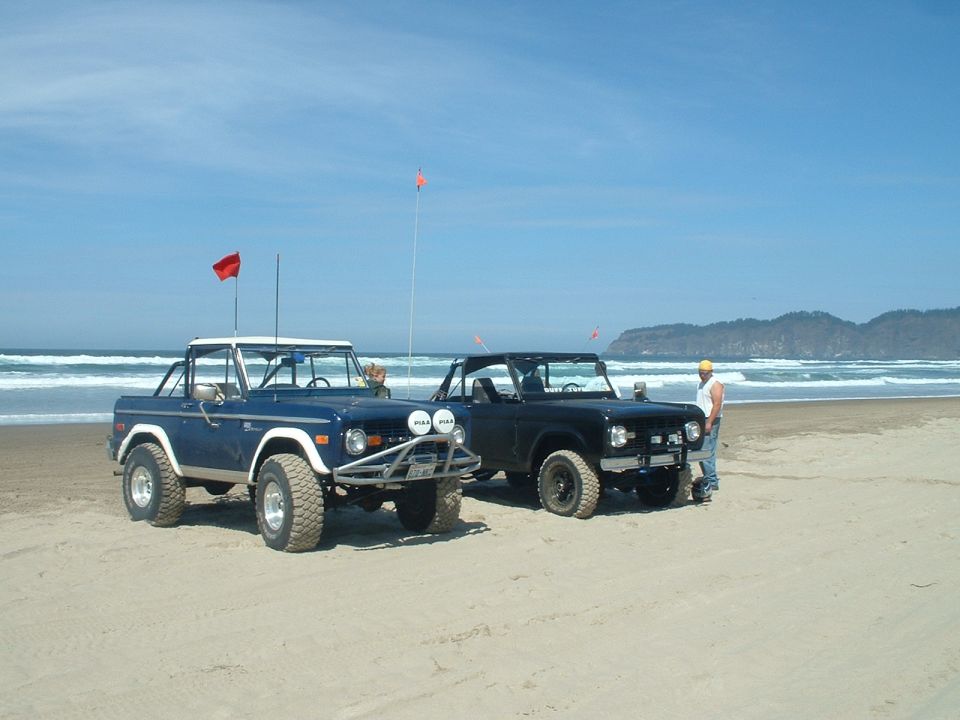Two early Ford Broncos parked on a beach, one blue and one dark, orange flags and surf behind