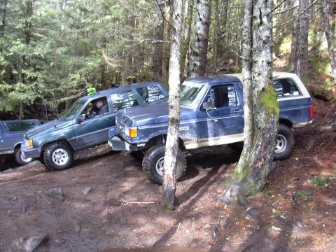 1991 Ford Bronco - Multiple off-road vehicles are seen on a challenging, muddy trail surrounded by trees.