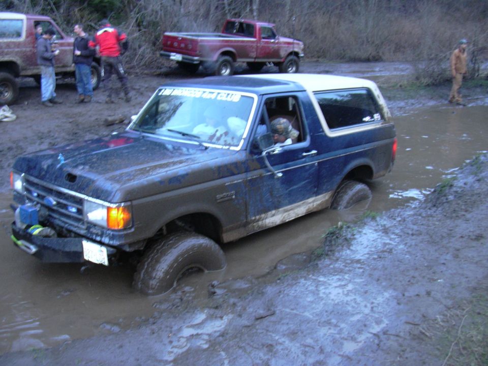 Muddy blue full-size Ford Bronco driving through a flooded rutted trail with people and a red truck behind