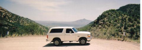 1996 Ford Bronco - This 1996 Ford Bronco is seen from the side, parked on a dirt road with a scenic mountain backdrop.