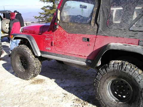 1996 Ford Bronco - A red Jeep Wrangler is parked on a snowy surface with other vehicles and people nearby.