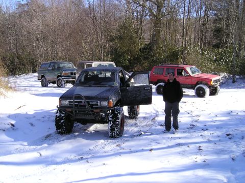 1996 Ford Bronco - Several modified off-road vehicles are parked in a snowy, wooded area with a person standing by one of them.