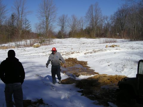 1996 Ford Bronco - Two people are in a snowy, wooded area with a muddy patch in the foreground. Part of an off-road vehicle is visible on the right.