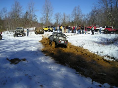1996 Ford Bronco - Off-road vehicles navigate a muddy track in a snowy environment, observed by a group of onlookers.