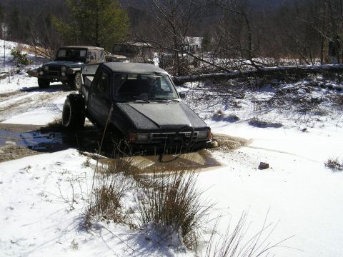 1996 Ford Bronco - Two off-road vehicles navigate a muddy, snow-covered trail in a wooded area.