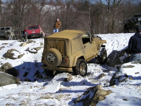 1996 Ford Bronco - Several off-road vehicles tackle a challenging snowy terrain with large rocks and trees in the background.
