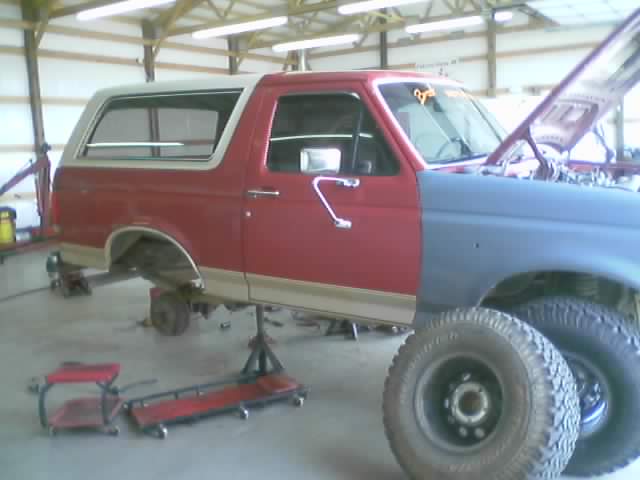 Red and white full-size SUV in garage with gray primer front fender, raised body, and removed tire on floor.
