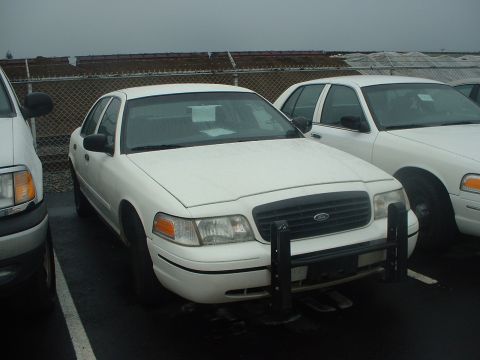 1999 Ford Crown Victoria - A white sedan with a push bar is parked in a lot next to another similar vehicle.