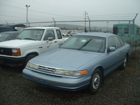 1999 Ford Crown Victoria - A light blue 1999 Ford Crown Victoria is parked next to a white pickup truck under an overcast sky.
